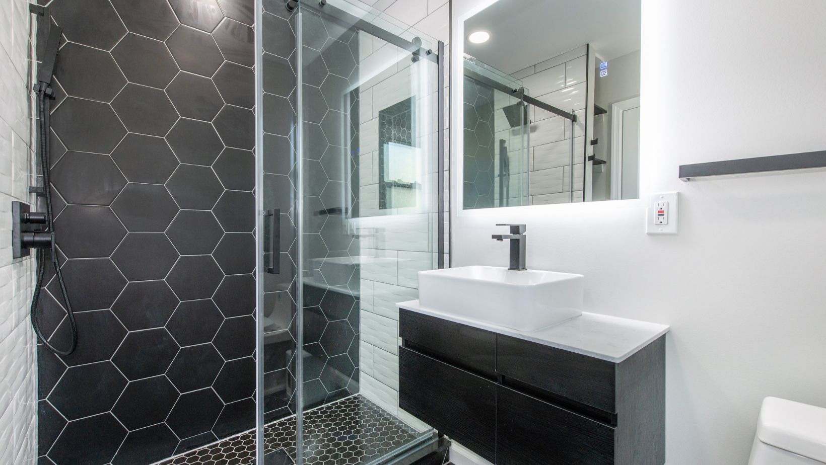 Bathroom with a dark floating vanity, white vessel sink, black fixtures, illuminated mirror, and glass shower with black hexagonal and white subway tiles.